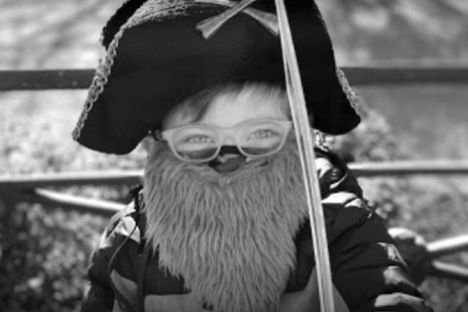 Child in pirate costume with hat, fake beard, and sword, sitting outdoors, in black and white.
